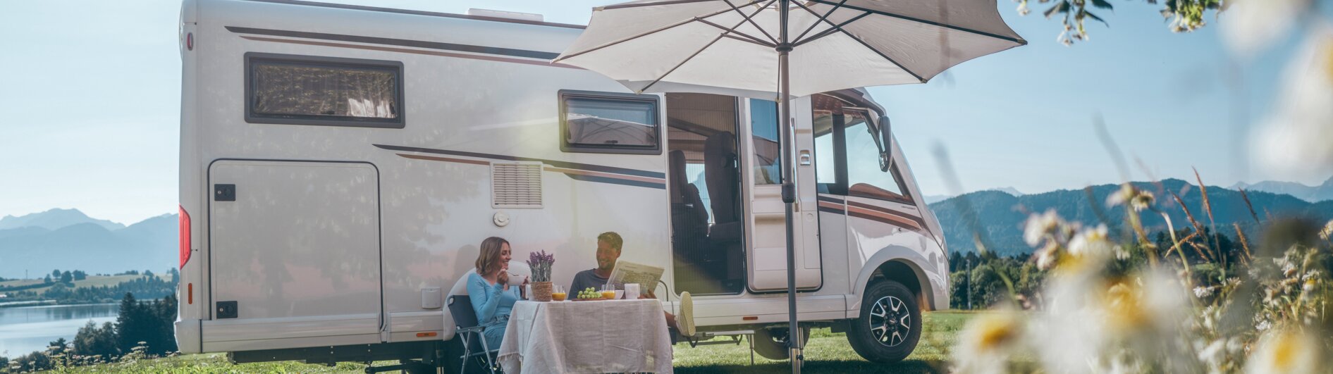 A man and a woman sitting in front of their campervan under a sunshade, drinking coffee.